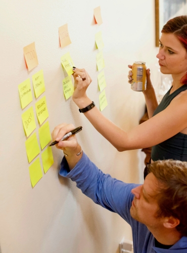 A woman writes on a yellow sticky note on a wall while a man sitting on the floor writes on another sticky note, both engaged in a brainstorming session.