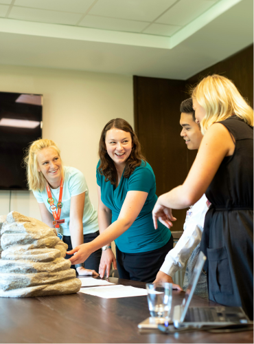 A group of four people discuss a stone object on a table in a conference room, engaged in a collaborative meeting.