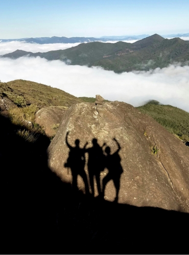 Four silhouetted figures pose on a rocky outcrop overlooking a mountainous landscape filled with clouds.