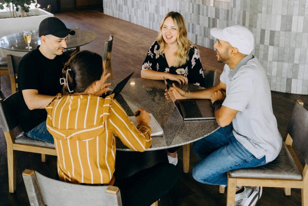 A group of four people, two men and two women, are seated around a circular granite table, engaging in a discussion with laptops and notebooks in a modern indoor setting.